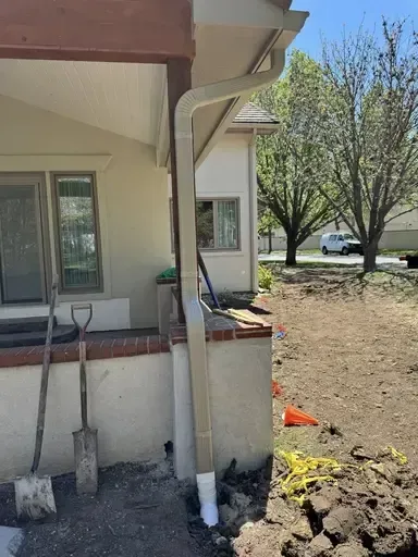Exterior view of a house with a gutter line running underground from downspout, tools, and dirt on a sunny day.