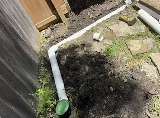 White PVC pipes laid in a trench along a wooden fence for an underground drain system, dirt and grass around.