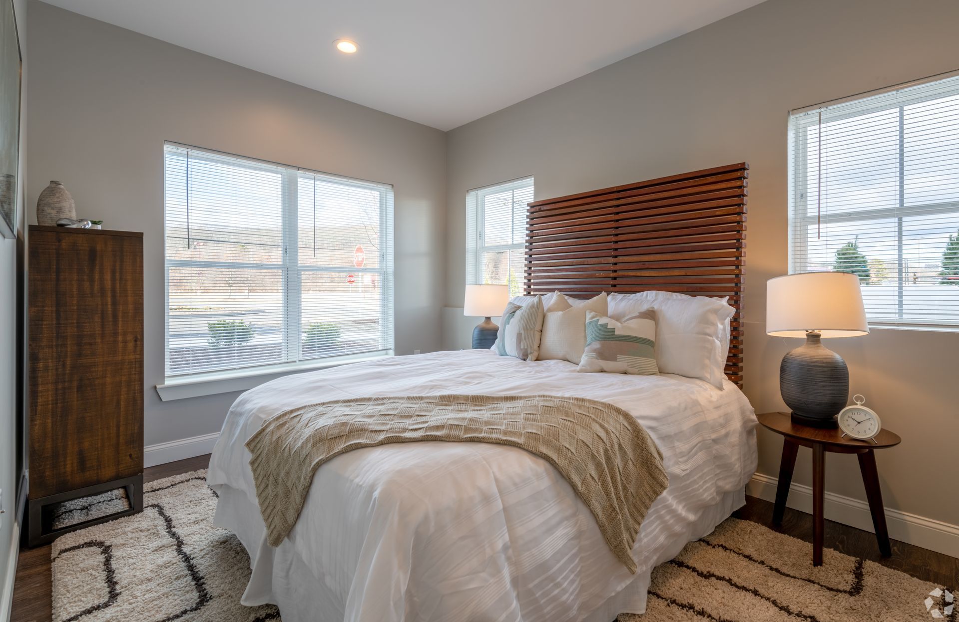 a bedroom with a bed , dresser , lamps and two windows .