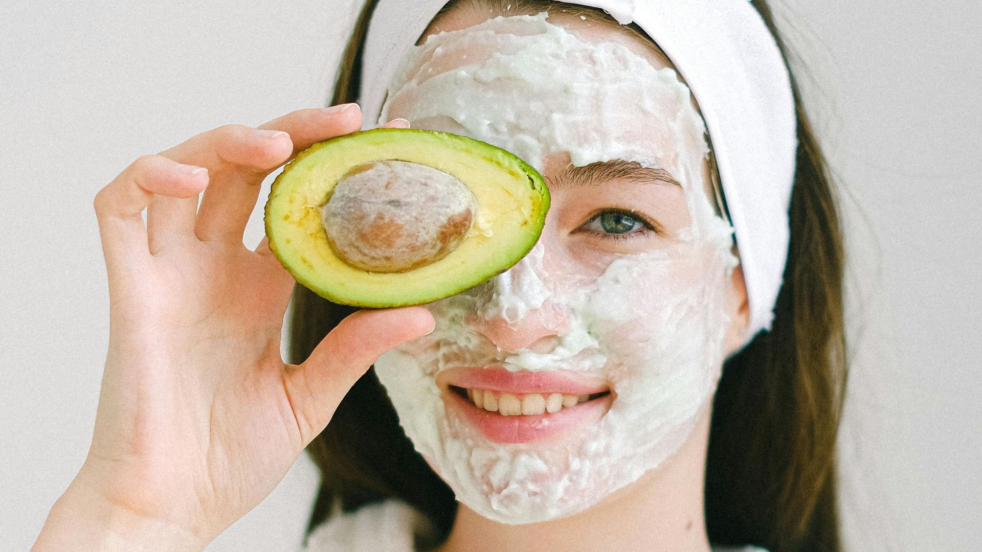 A woman with an avocado mask on her face is holding a slice of avocado in front of her eye.