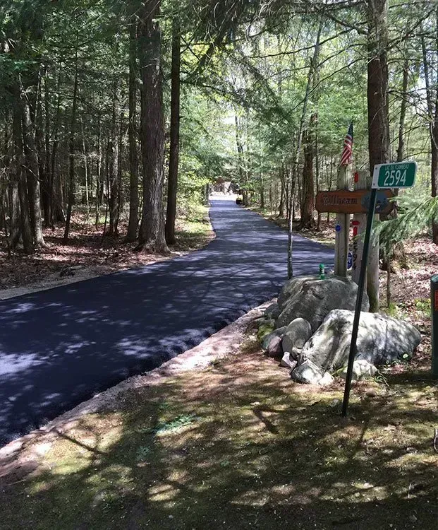A paved driveway leads into a wooded area, past a signpost with a green house number sign and large rocks in the foreground.