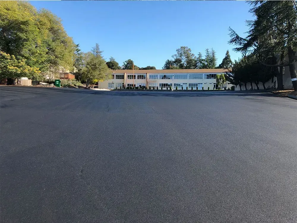 A wide, freshly paved dark asphalt parking lot leading up to a beige, multi-story commercial building under a clear sky.