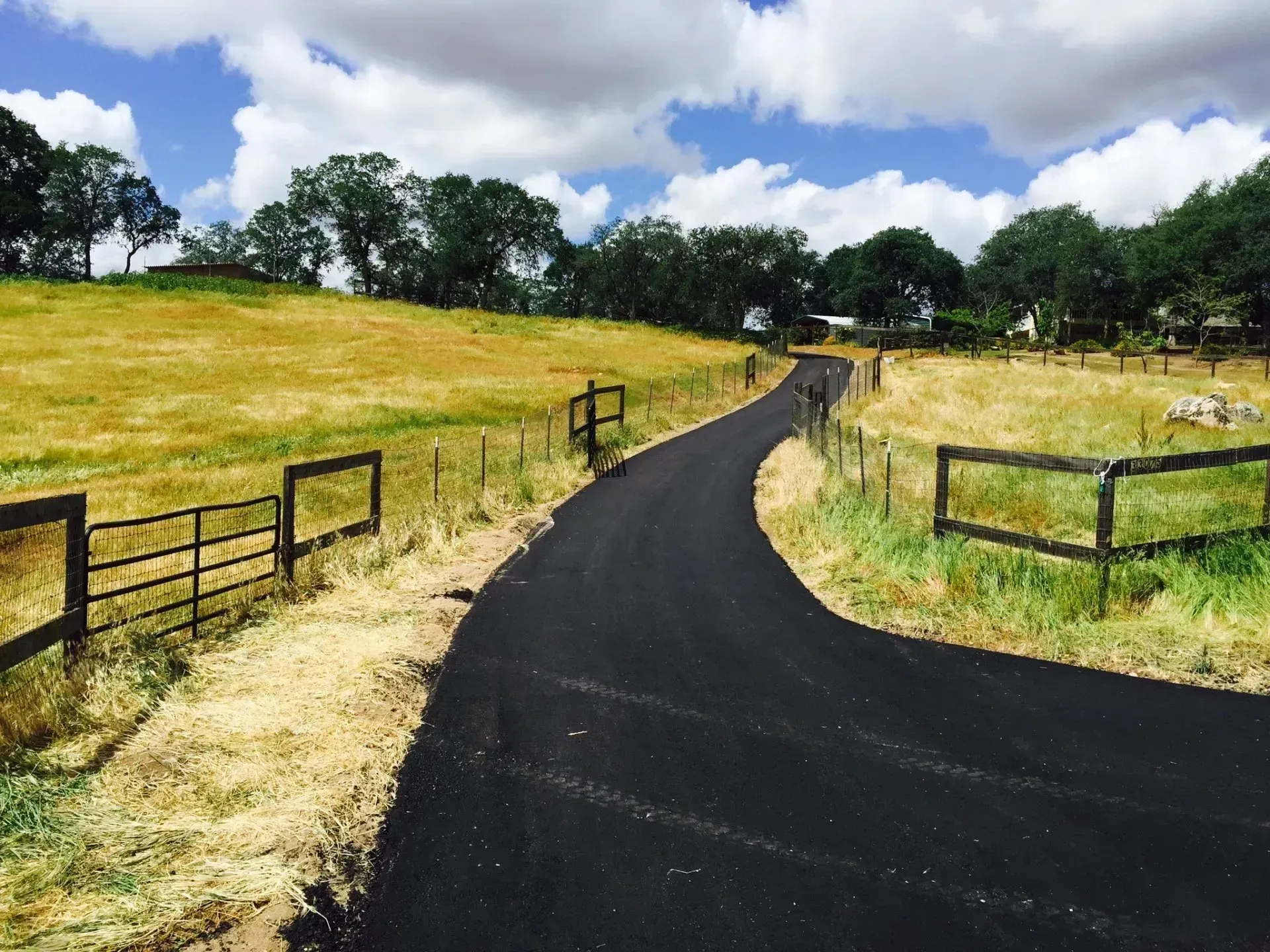 A paved driveway curves through a rural landscape with dry, golden fields bordered by wooden fences under a cloudy sky.