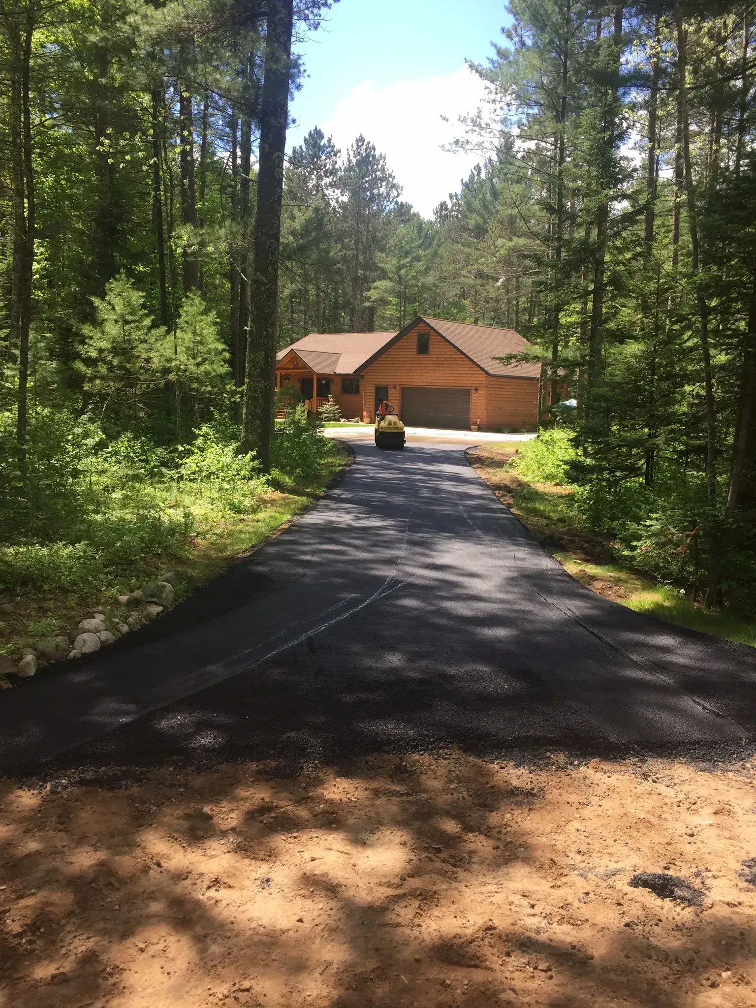 A newly paved asphalt driveway leads through a wooded area toward a brown, one-story house.