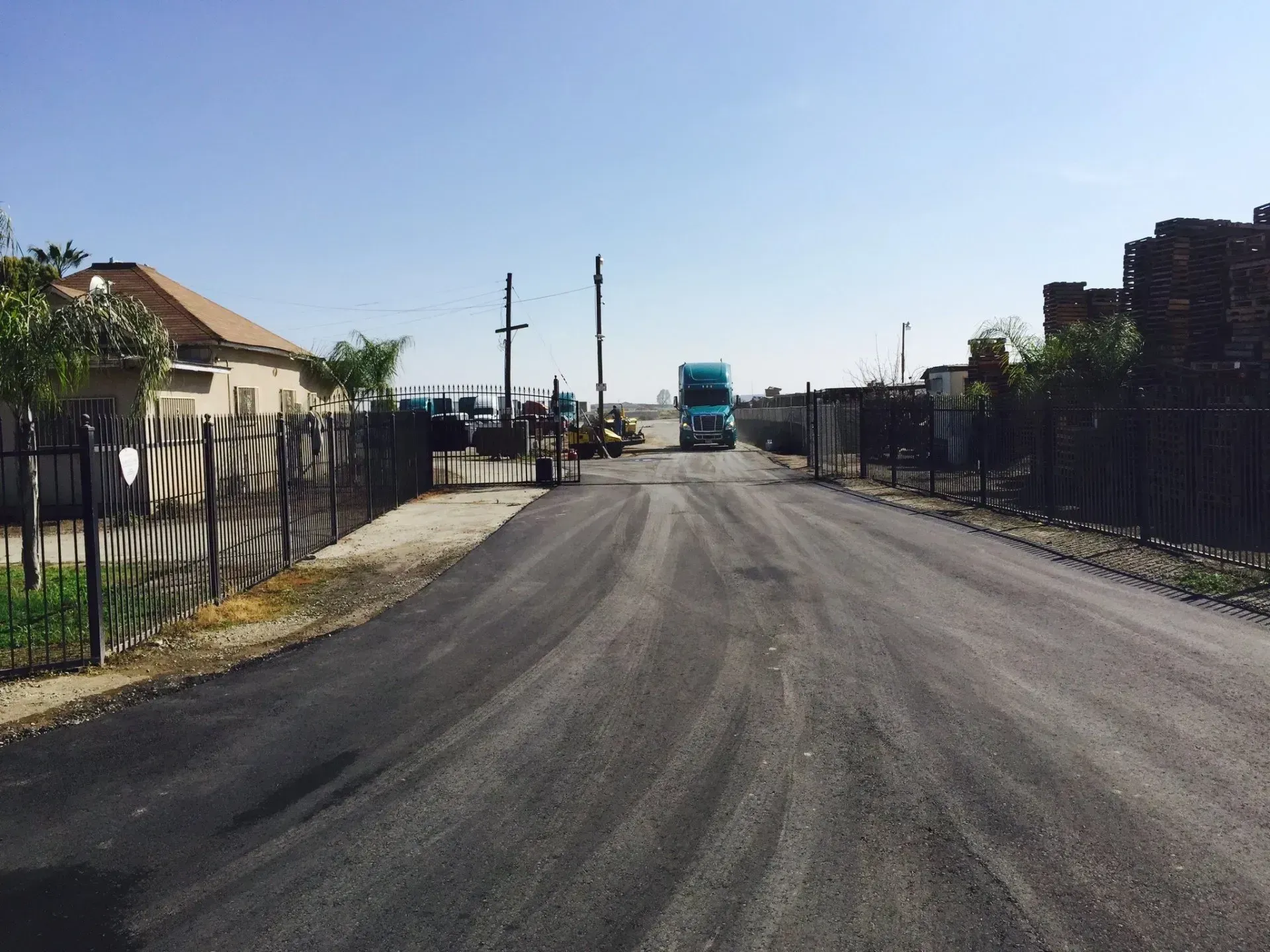 A paved road leads toward a light blue semi-truck parked in the distance between residential and industrial buildings.