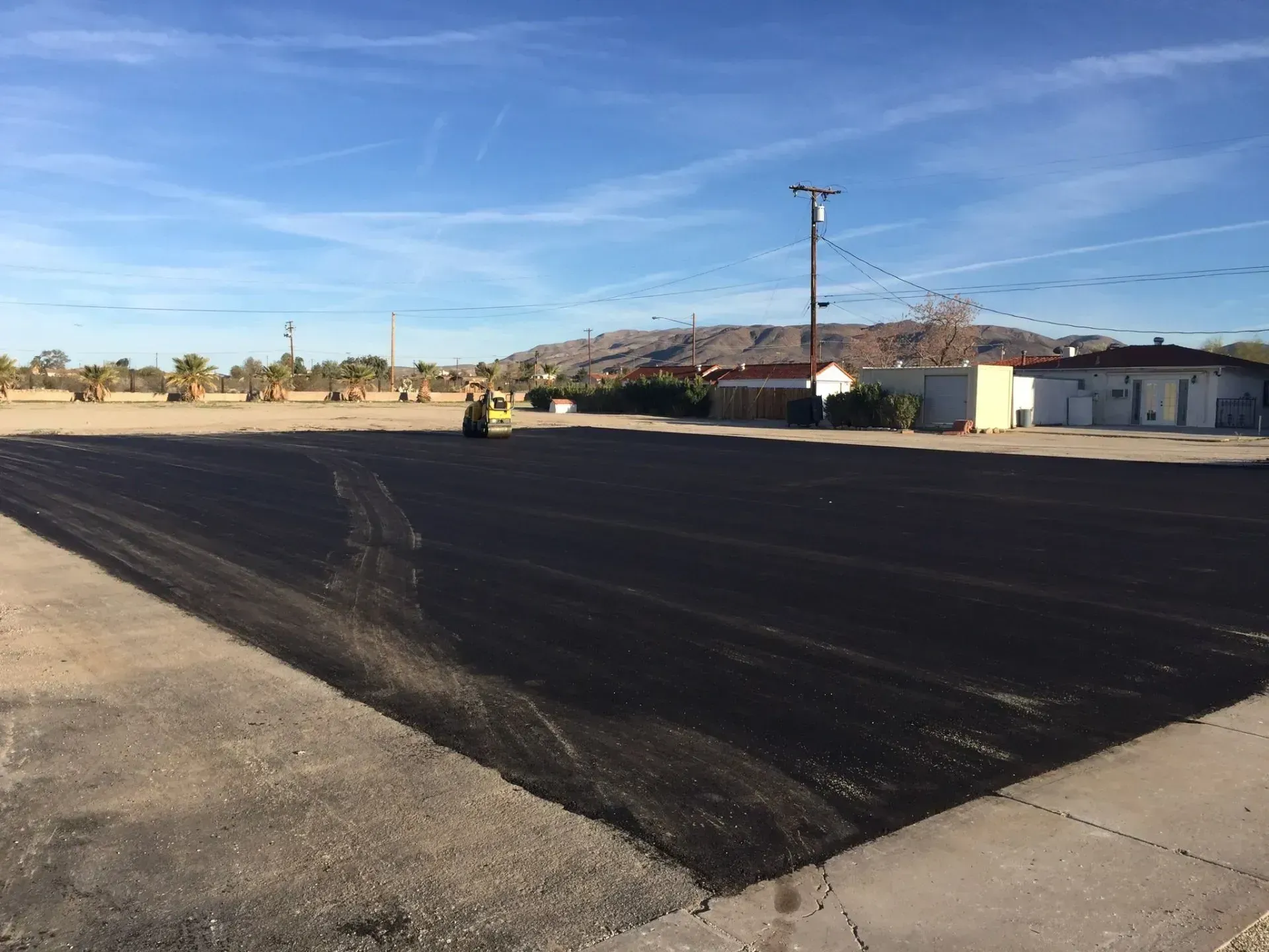 A yellow steamroller compacts fresh, dark asphalt on a large lot under a clear blue sky, with buildings in the distance.