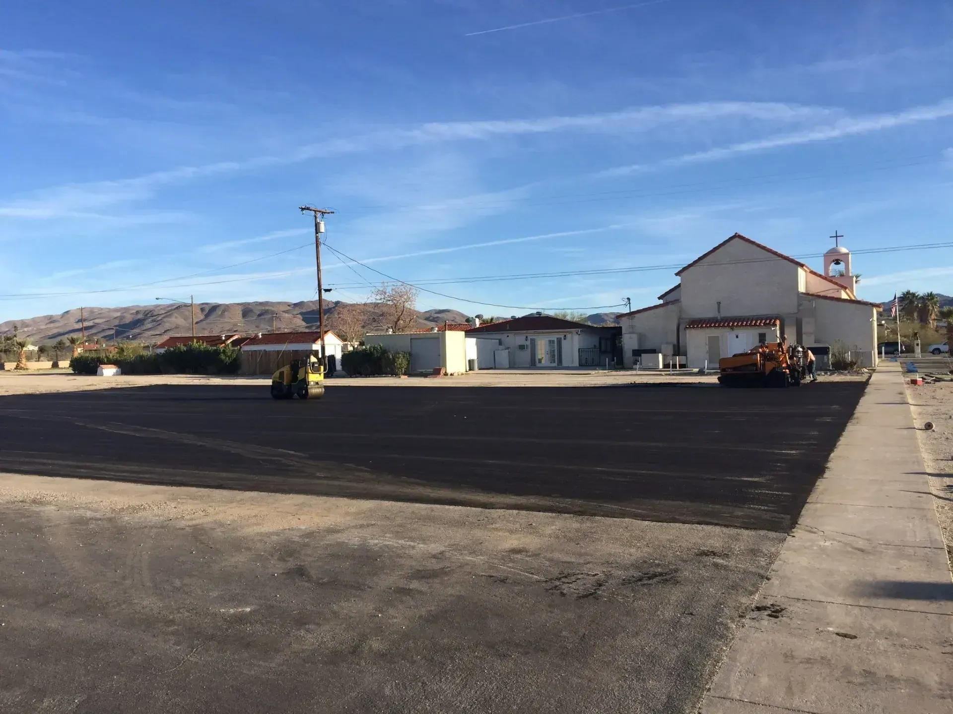 Construction workers use heavy rollers to pave a large asphalt parking lot in front of a white church building.