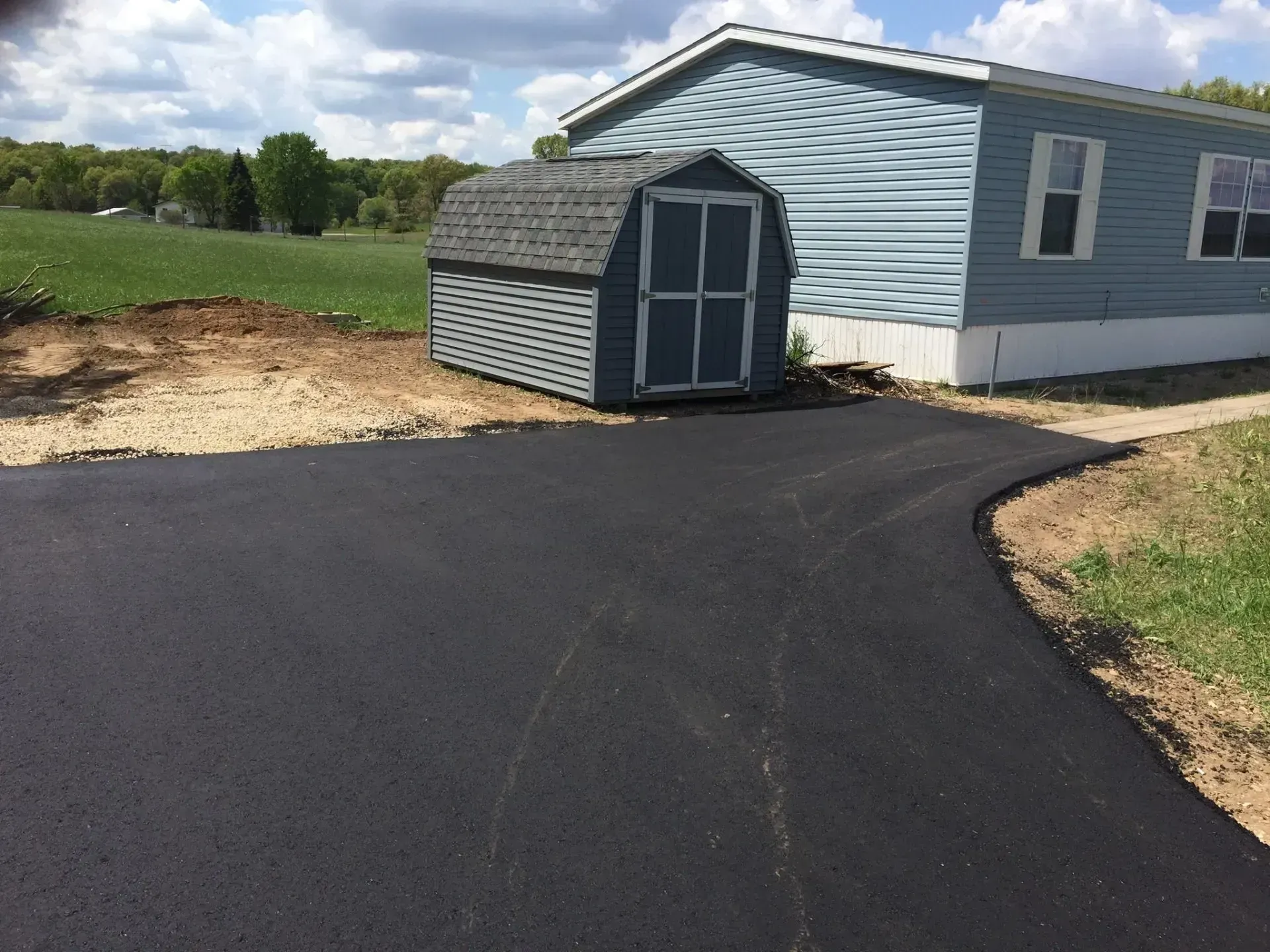 A newly paved asphalt driveway leads toward a blue storage shed next to a light blue mobile home in a grassy yard.