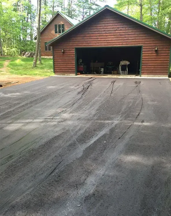 A wooden garage with an open door stands behind a newly paved asphalt driveway featuring prominent tire tracks.