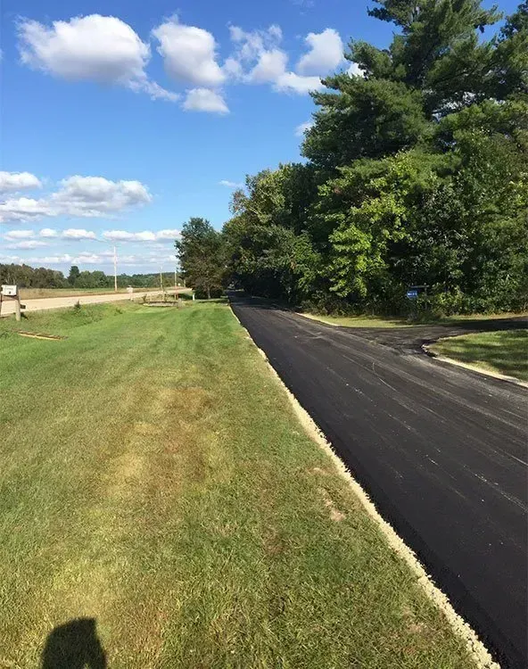 A freshly paved black asphalt road runs alongside a bright green grassy field under a sunny blue sky with scattered clouds.