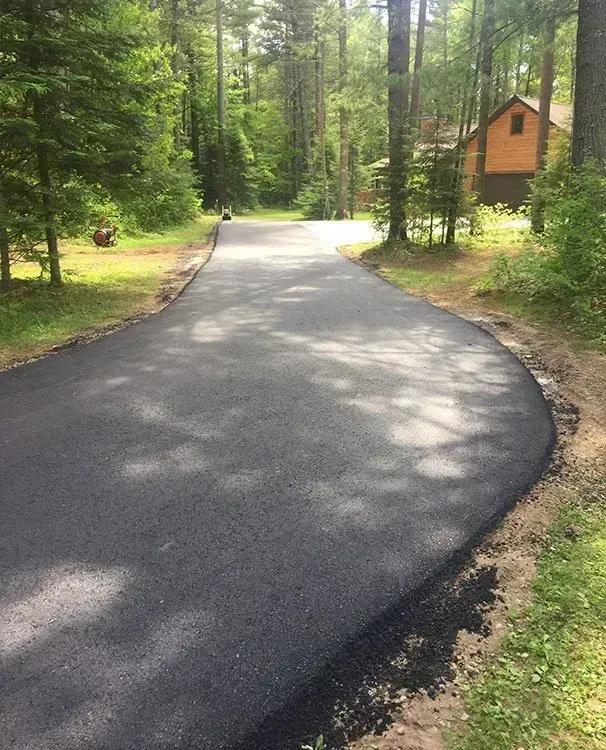 A freshly paved black asphalt driveway curves through a sunlit wooded area toward a house in the distance.