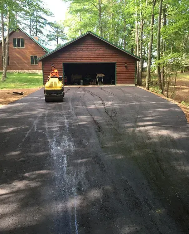 A yellow steamroller compacts fresh black asphalt on a driveway in front of a wooden garage and a house in the woods.
