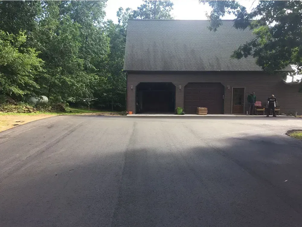A newly paved asphalt driveway leads to a large, two-bay garage nestled in a wooded area.