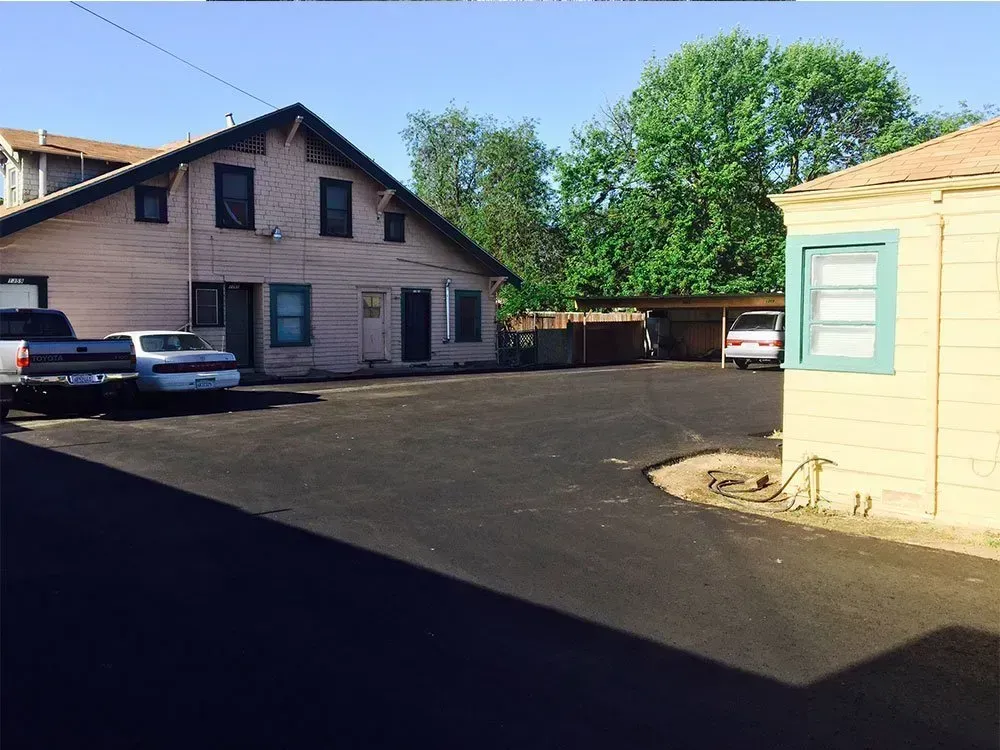A paved lot between a two-story pink residential building and a smaller tan structure with a light blue window frame.