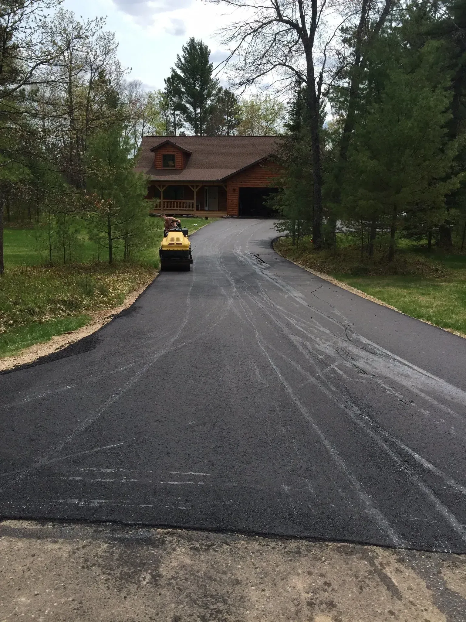 A yellow steamroller compacts fresh, dark asphalt on a driveway leading to a house surrounded by trees.