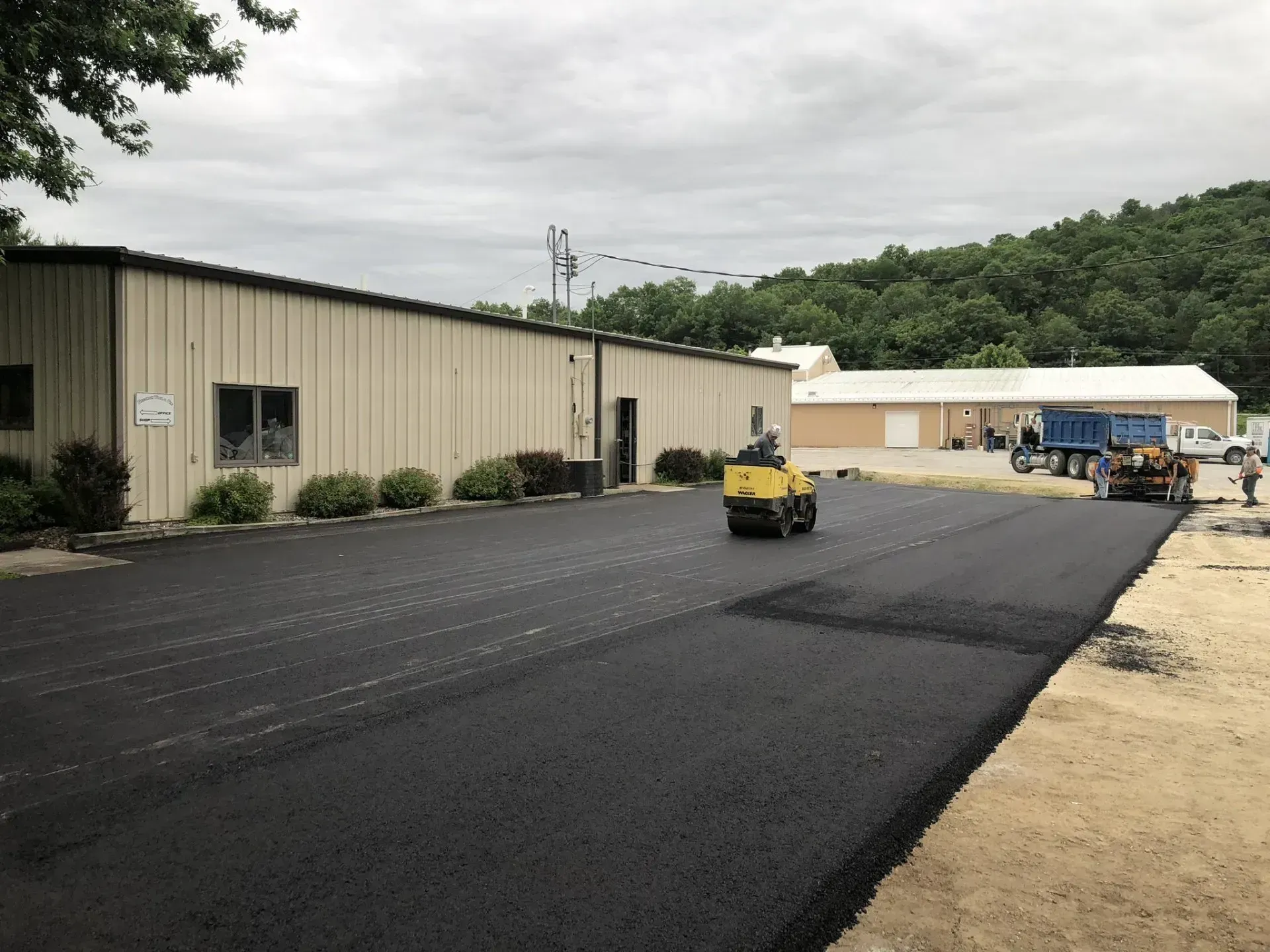 A yellow asphalt roller compacts a freshly paved black parking lot next to a tan commercial building.