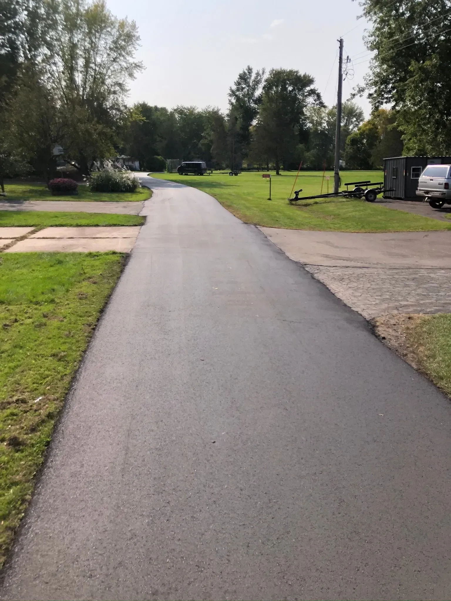 A freshly paved black asphalt driveway stretches through a grassy area toward trees under a clear sky.