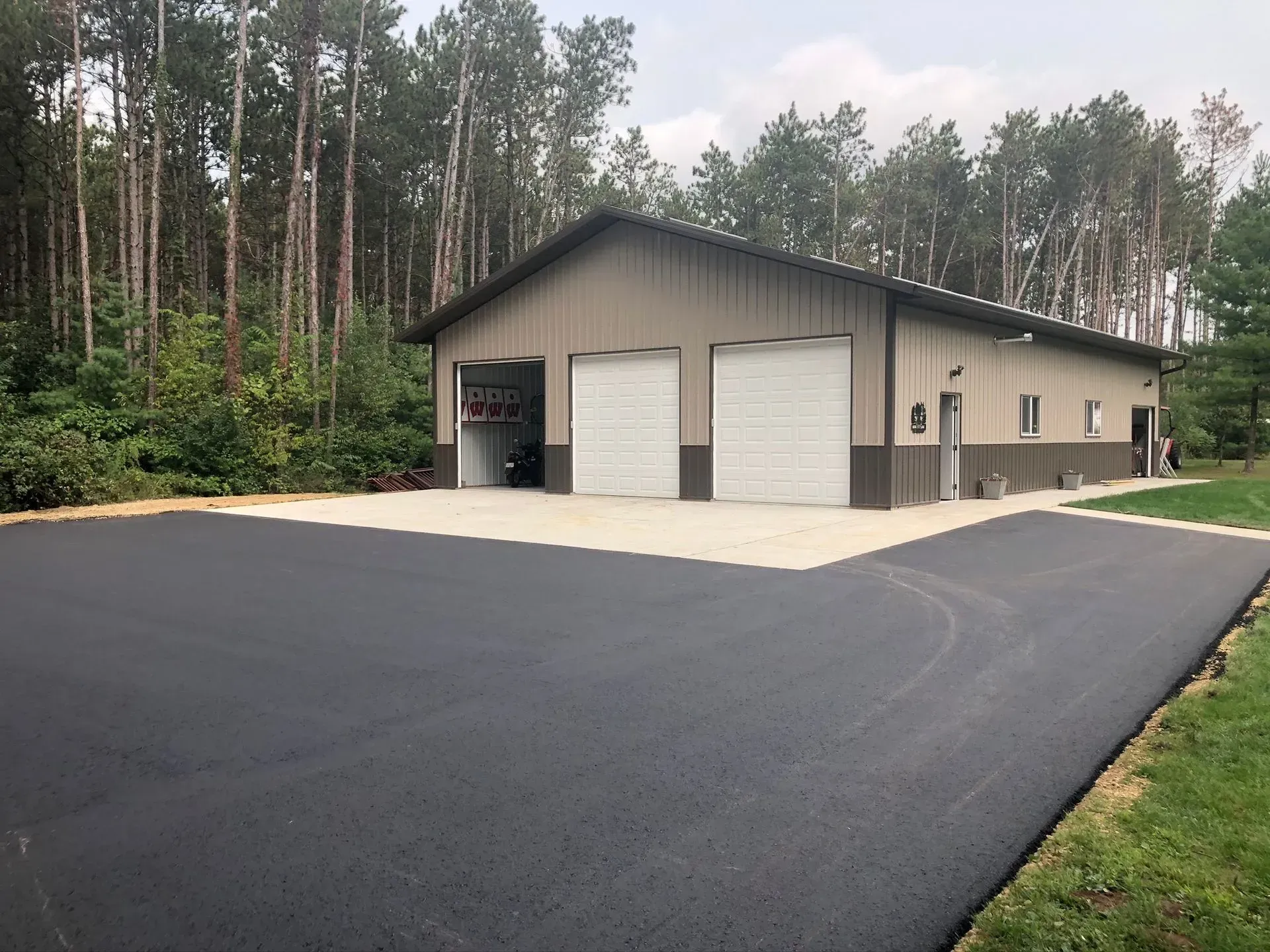 A tan, metal-sided building with two white garage doors, set on a large, freshly paved asphalt driveway near a forest.