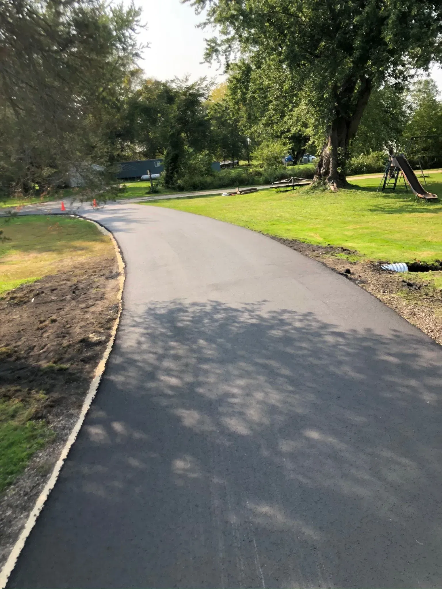A newly paved asphalt driveway curves through a grassy yard toward trees on a sunny day.