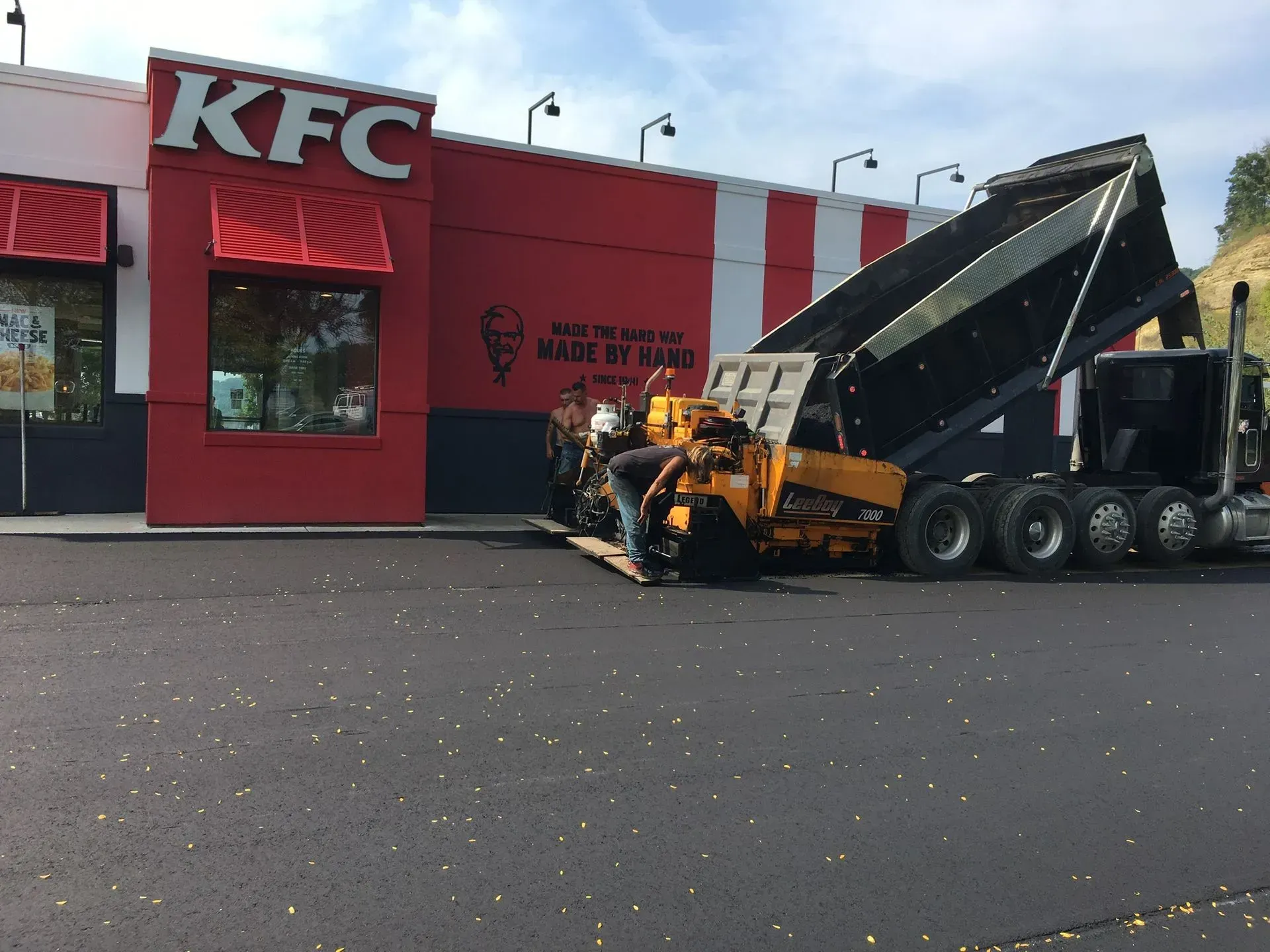 A yellow construction paving machine works on a freshly laid black asphalt parking lot in front of a KFC restaurant.