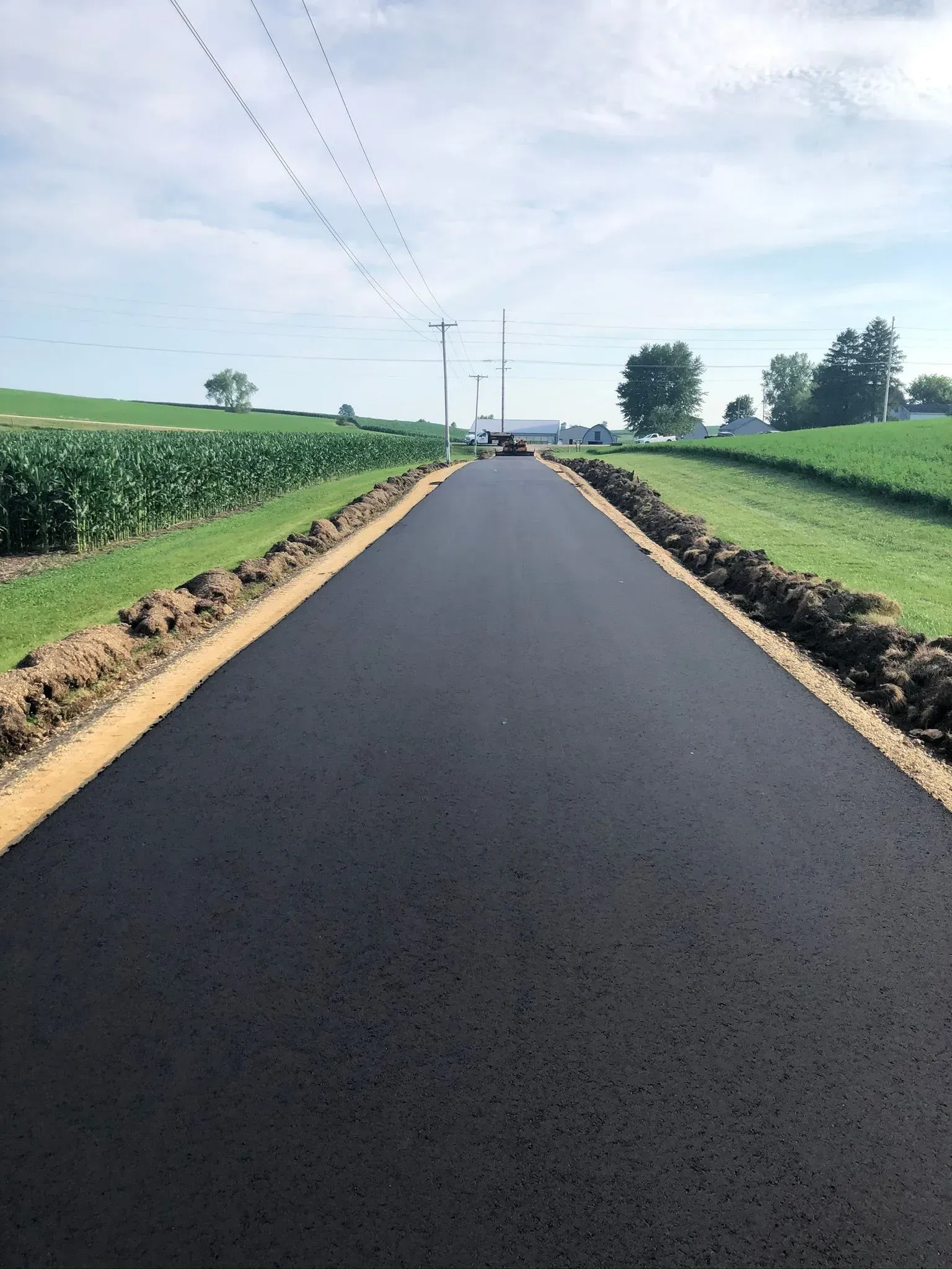A newly paved, dark asphalt road stretches into the distance, flanked by green fields and fresh soil shoulders.