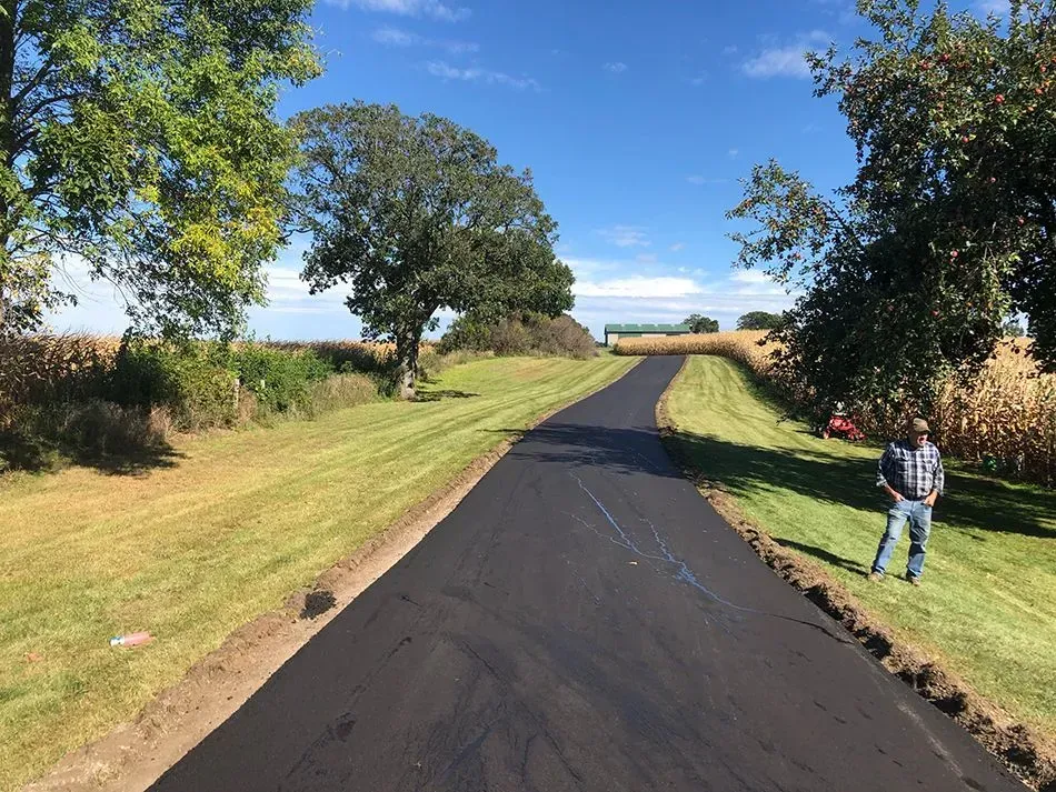 A newly paved asphalt driveway stretches through a grassy field toward a distant farm building under a clear blue sky.