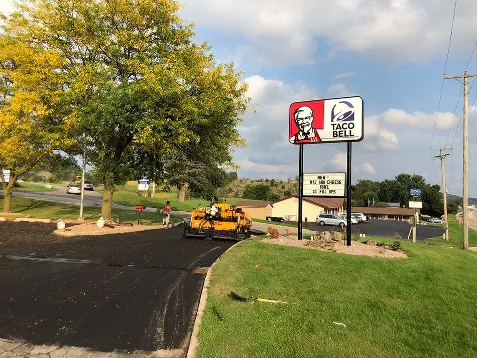 A KFC and Taco Bell sign stands next to an area where road construction equipment is laying fresh asphalt.
