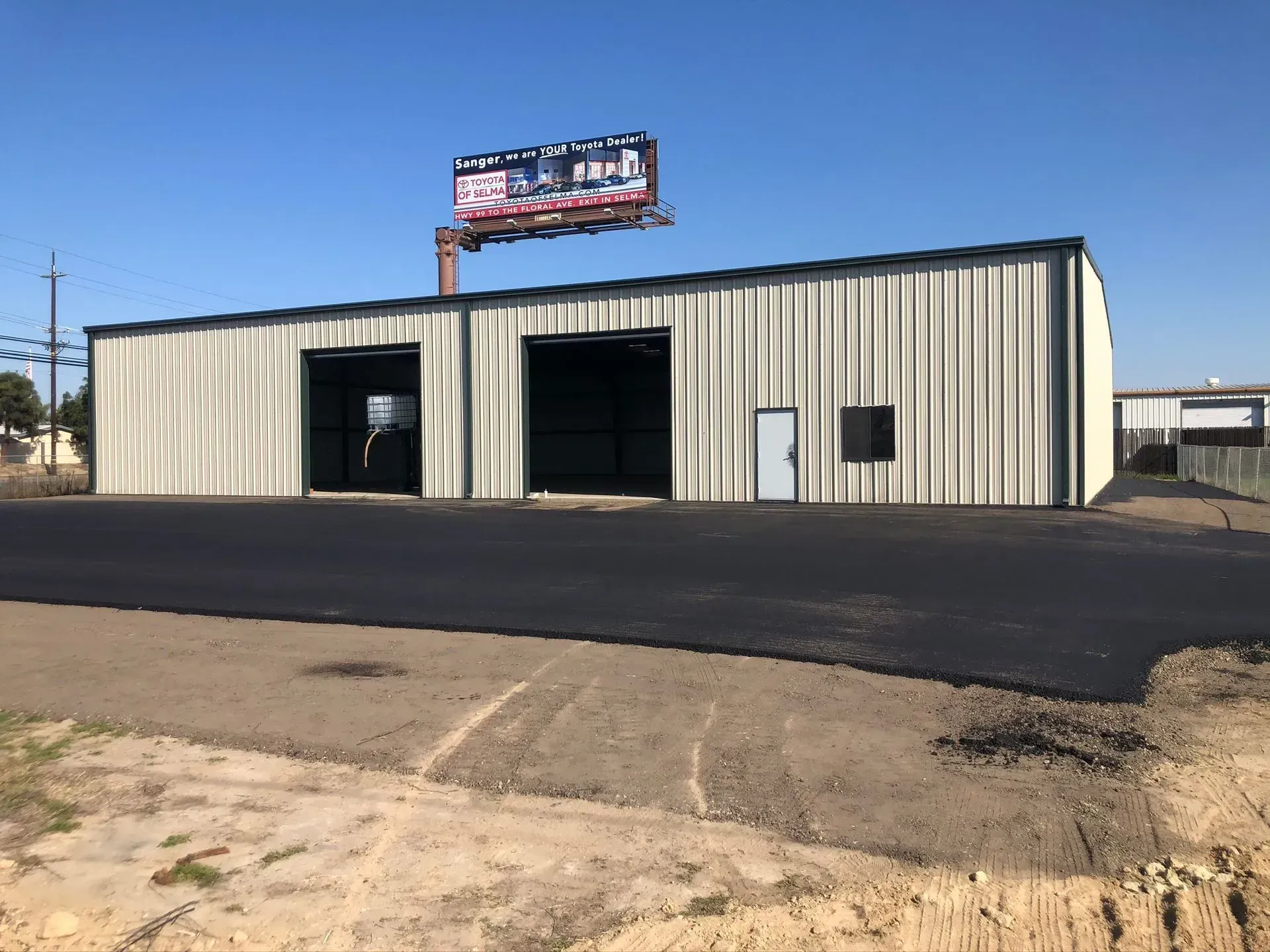 A beige metal warehouse with two open bays, a side door, and a billboard on the roof, viewed from a freshly paved lot.