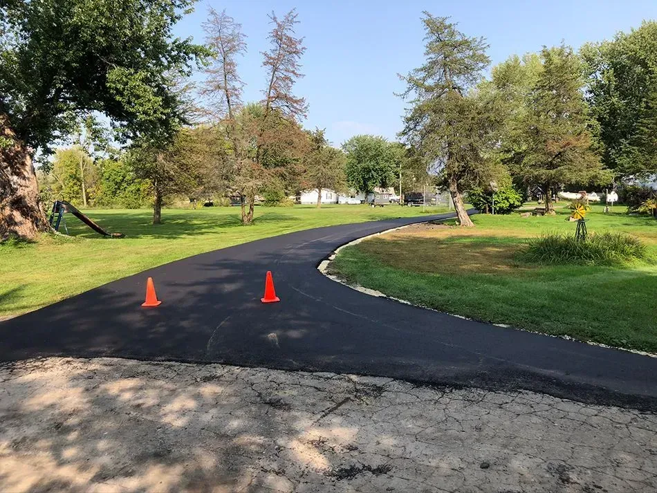 A freshly paved black asphalt driveway curves through a green, grassy yard, marked by two small orange traffic cones.