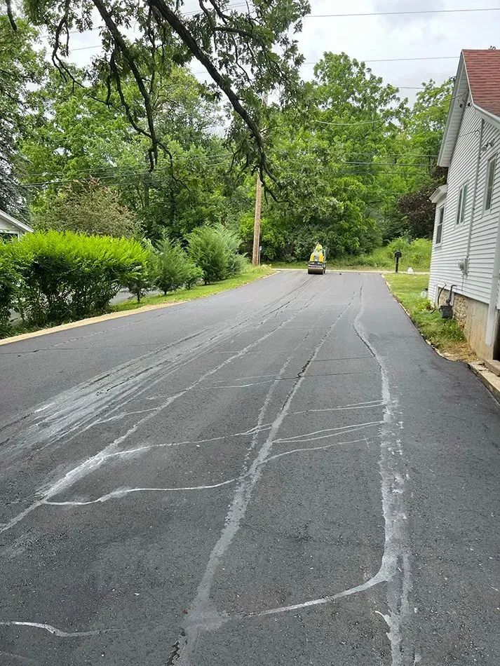 A freshly paved asphalt driveway with visible sealant lines, leading toward a house and a small road roller in the distance.