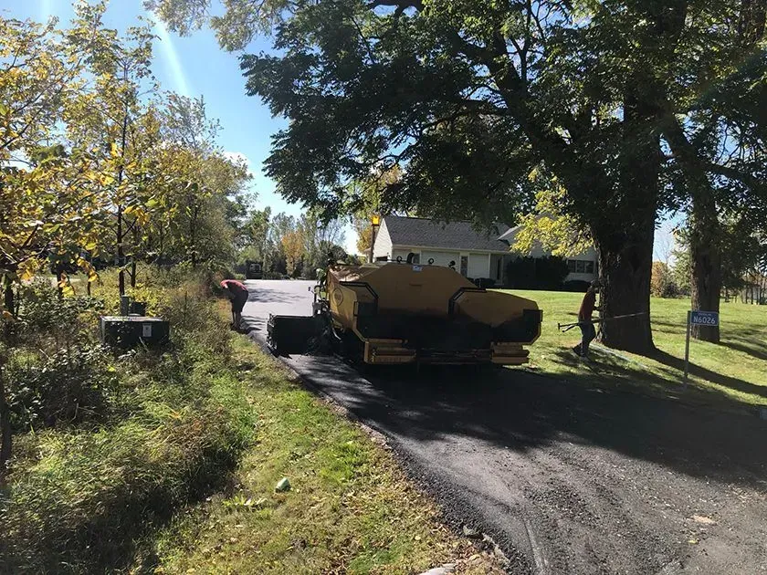 Workers operate a yellow paving machine to resurface a driveway on a sunny day with trees in the background.