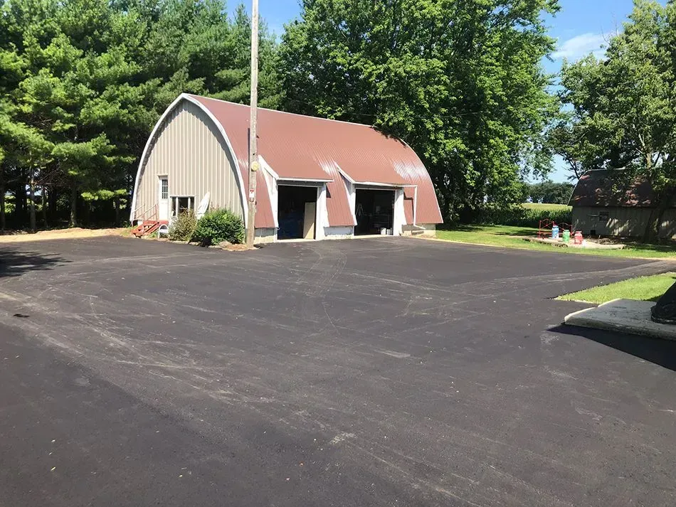 A tan, arched-roof building with two garage bays sits beside a large, freshly paved black asphalt parking lot.