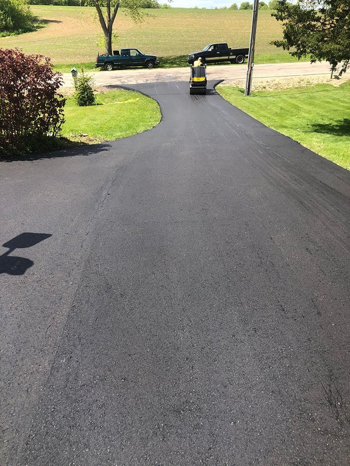 A freshly paved asphalt driveway curves toward a road with two parked pickup trucks in the background.