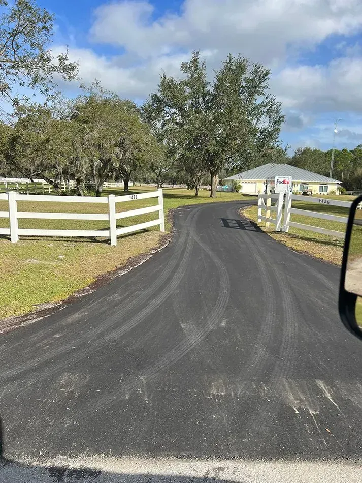 A black paved driveway leading to a house in the distance, framed by white wooden fences and trees under a blue sky.
