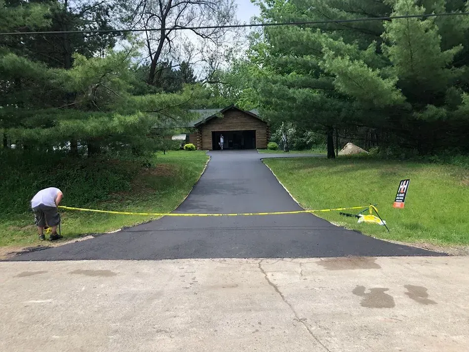 A worker stands by a freshly paved asphalt driveway marked with yellow caution tape leading to a wooden house.