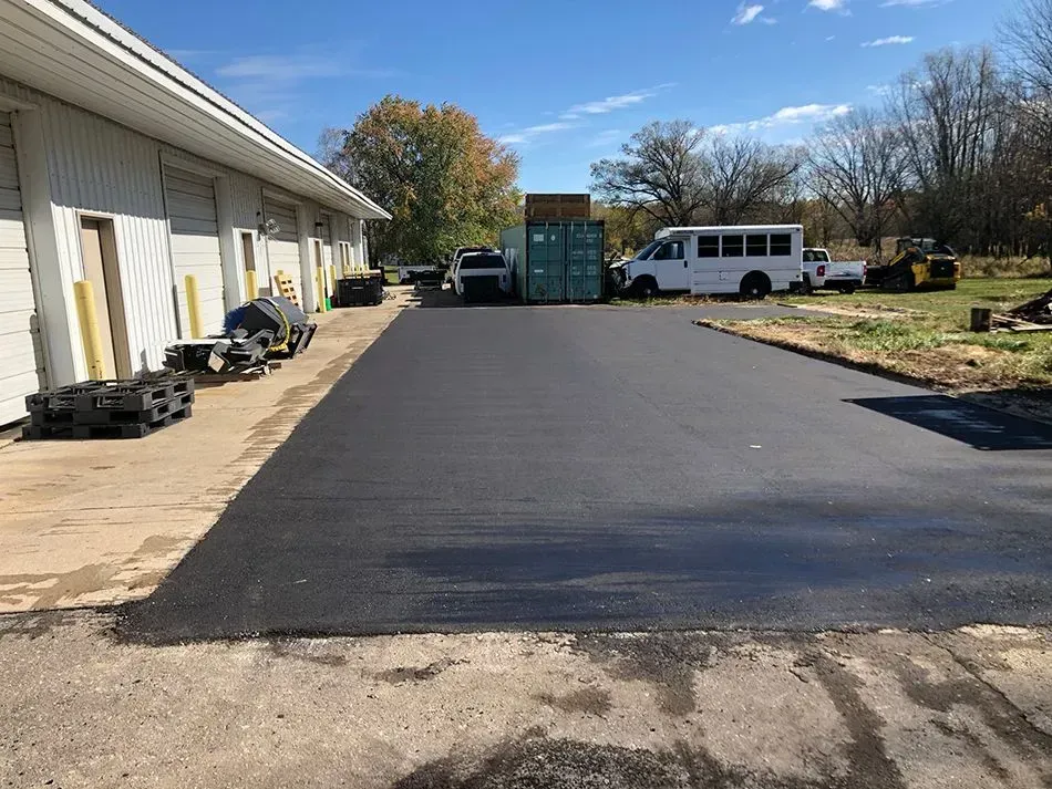 A newly paved asphalt driveway next to a warehouse building, with a white shuttle bus and storage container in the back.