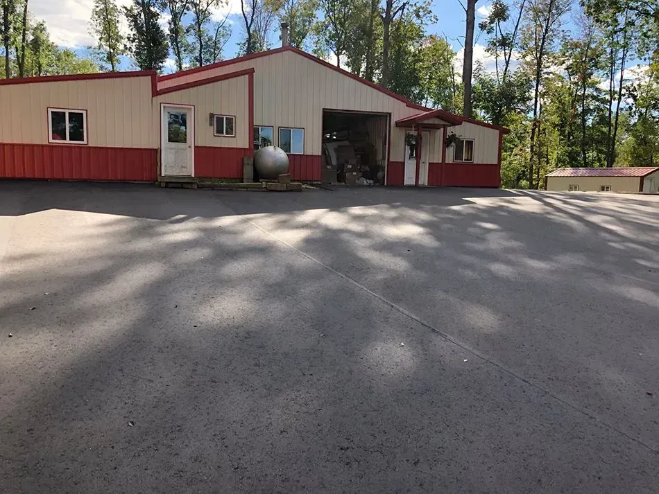 A wide view of a beige and red metal building with a large open garage door and a paved parking area, surrounded by trees.