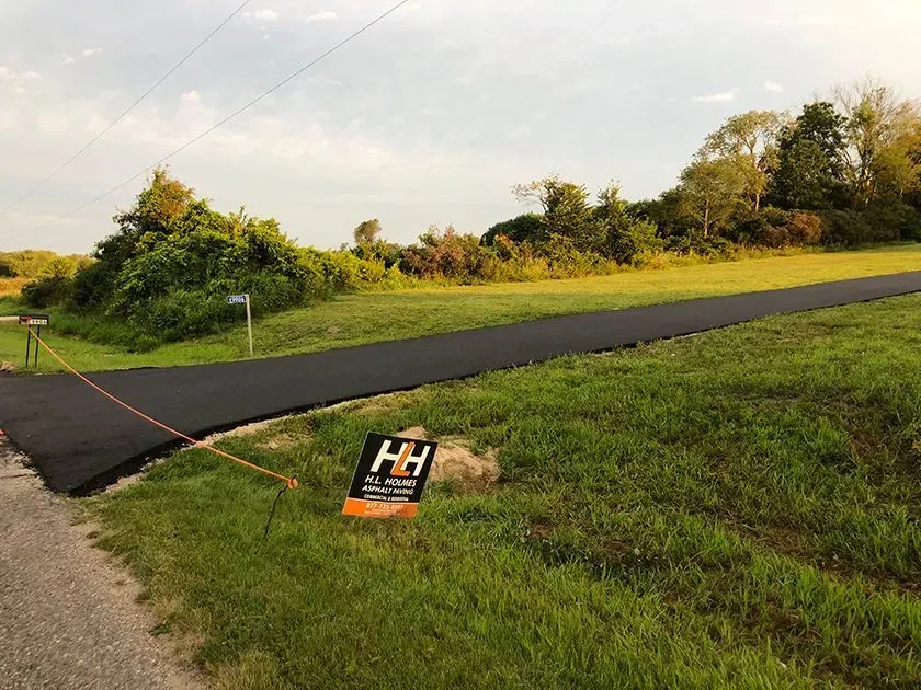 A newly paved black asphalt driveway leads into a grassy, undeveloped lot with a small H&H real estate sign in the foreground.