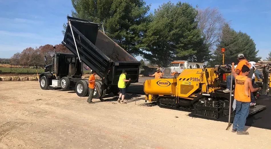 A dump truck is discharging material into a yellow asphalt paver while road workers operate the machinery outdoors.