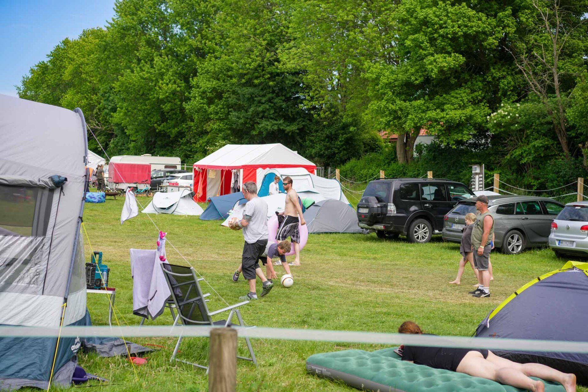 Campingplatz mit Zelten, Autos und Leuten, die auf dem Gras unter Bäumen Fußball spielen.