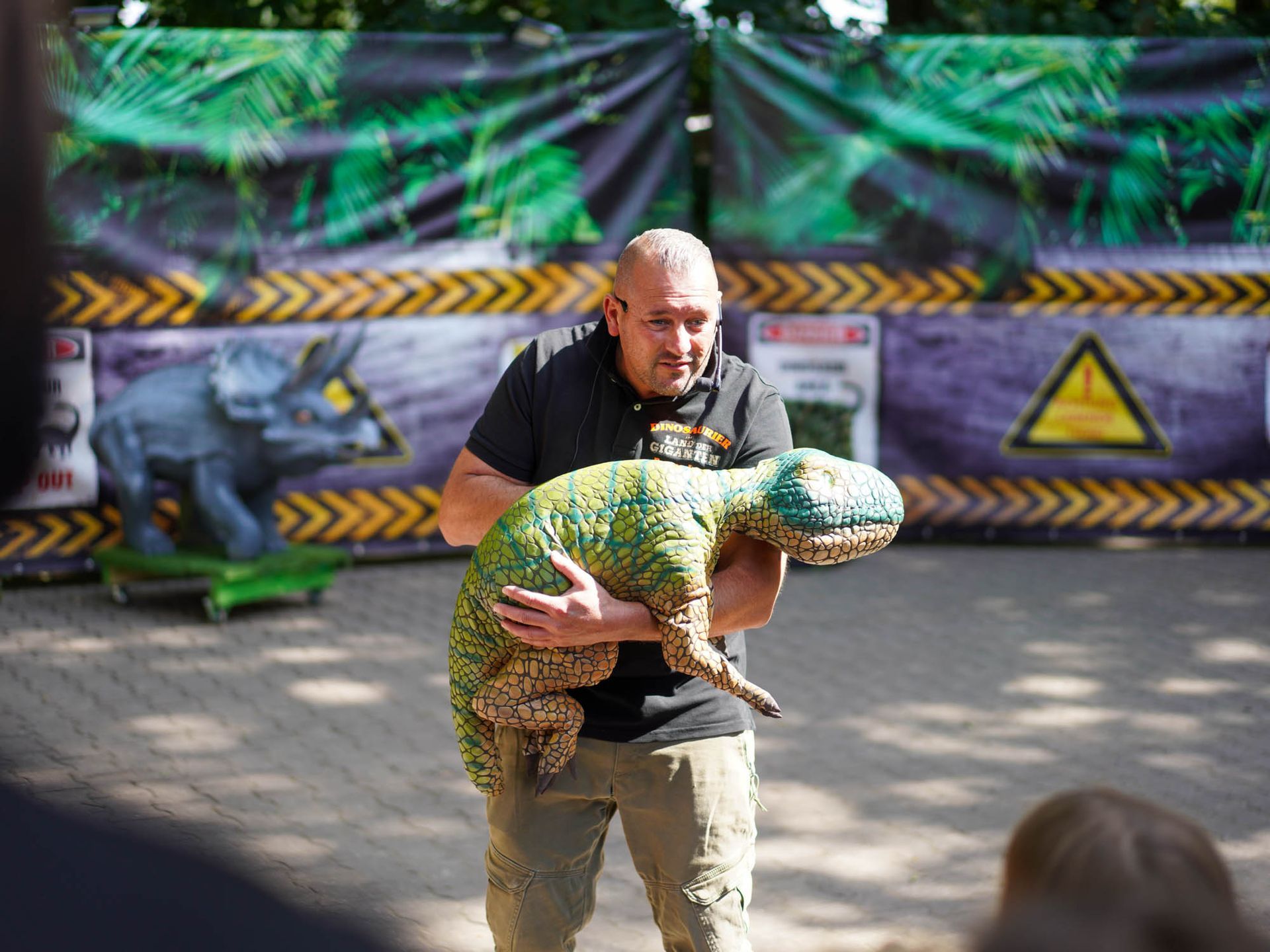 Mann hält eine kleine Dinosaurierpuppe vor einem Bühnenhintergrund mit Dinosauriermotiven.