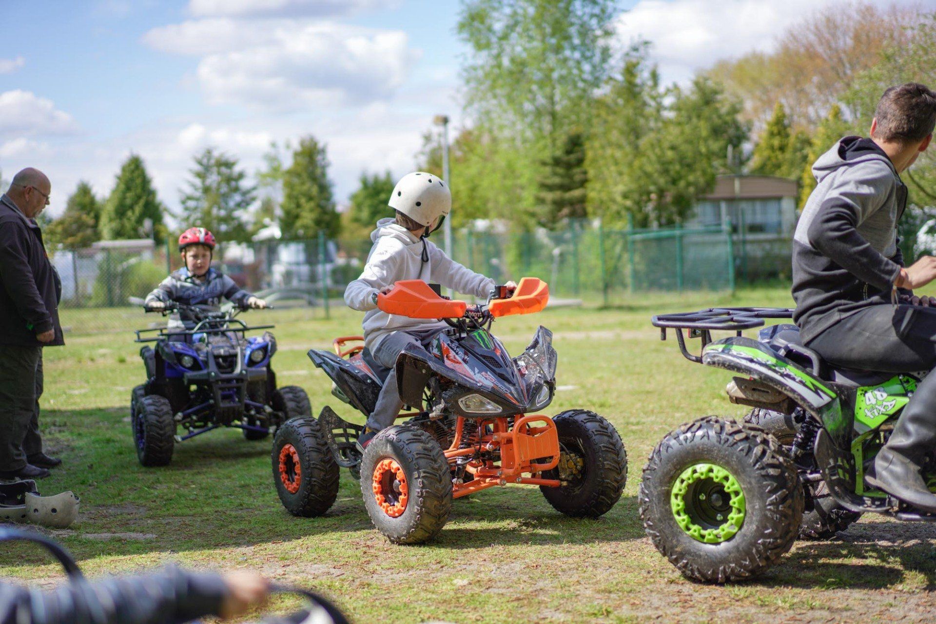 Kinder fahren mit Geländefahrzeugen auf Gras, tragen Helme und werden von Erwachsenen beaufsichtigt.