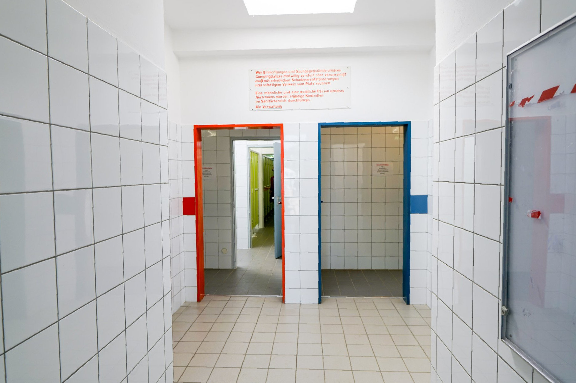 Flur mit weiß gefliesten Wänden, zwei Türen (rot und blau) und einem Whiteboard.