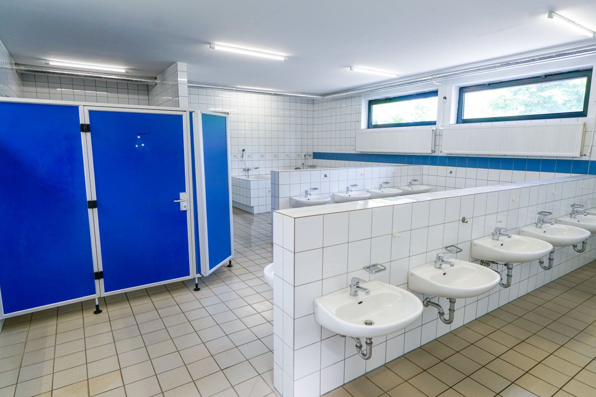 Blau und weiß geflieste öffentliche Toilette mit Waschbecken, Kabinen und Fenstern.