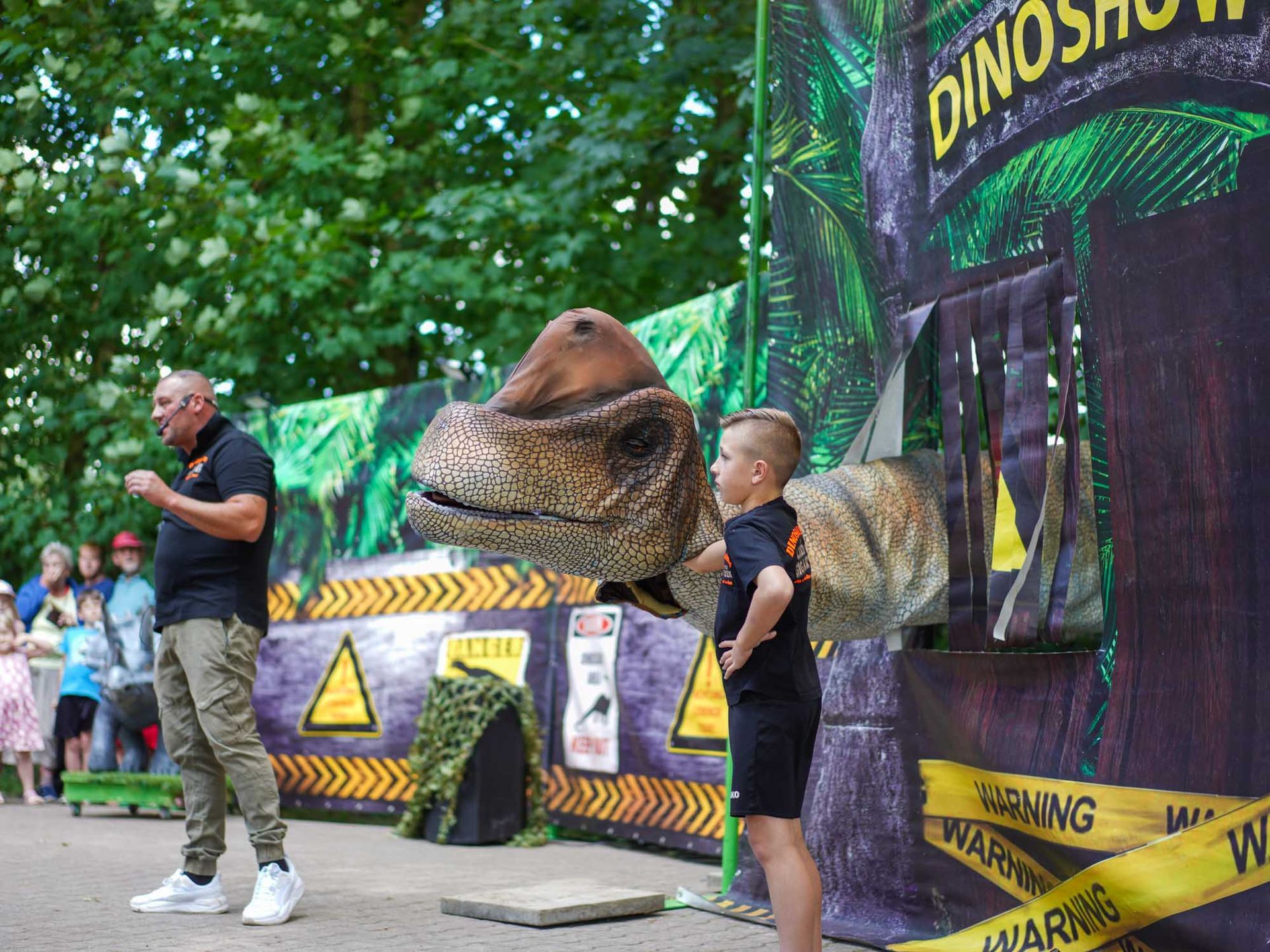 Mann und Junge mit einem Dinosaurier-Puppenkopf bei einer „Dino-Show“ mit Publikum im Freien.