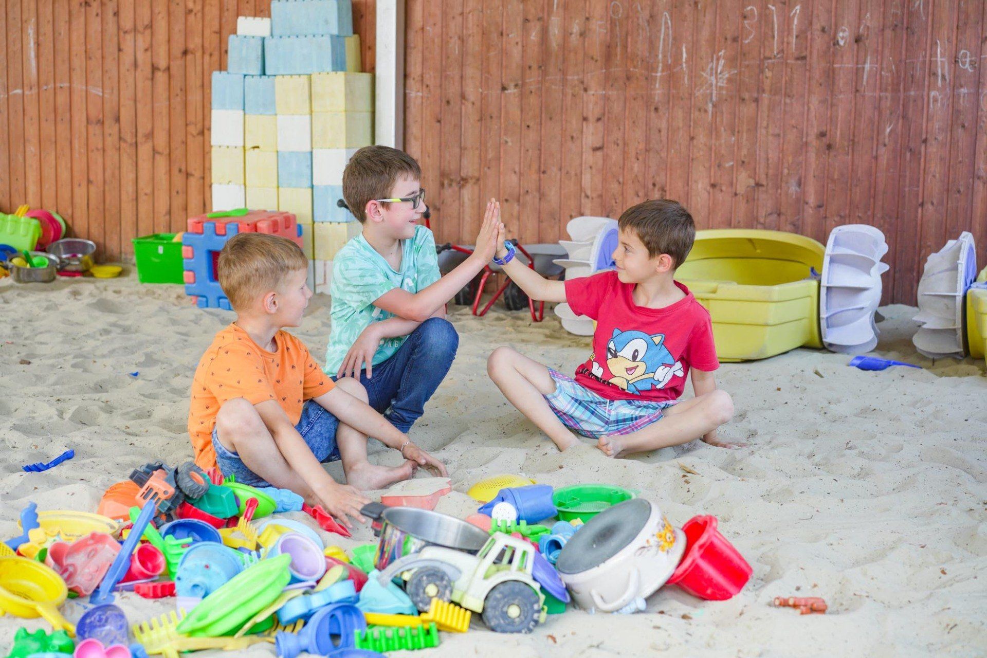 Drei Jungen geben sich in einem Sandkasten, umgeben von Spielzeug, ein High-Five. 