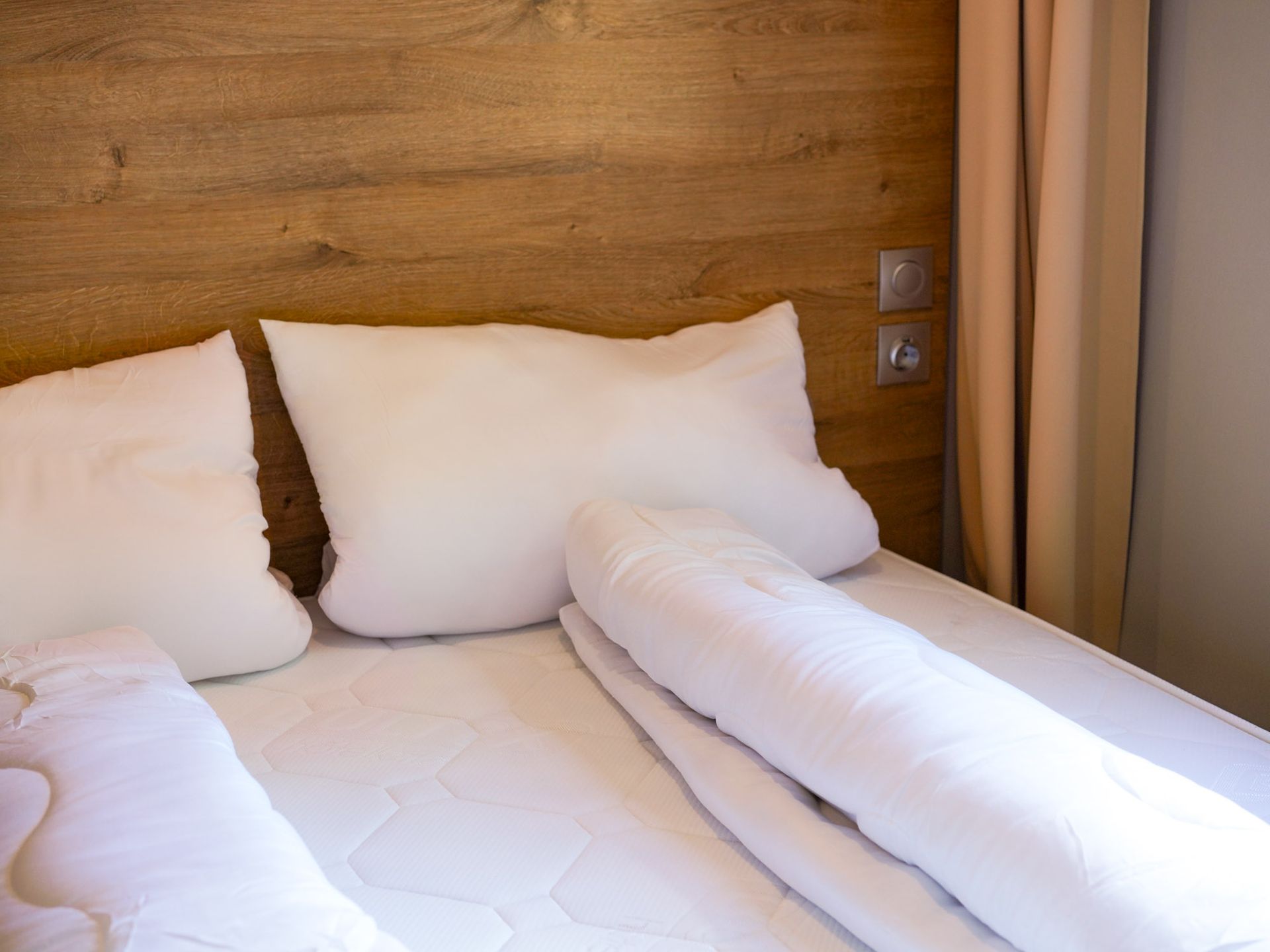 Weiße Bettwäsche auf einem Bett mit einem hölzernen Kopfteil, Kissen und Vorhängen in neutralen Farben.