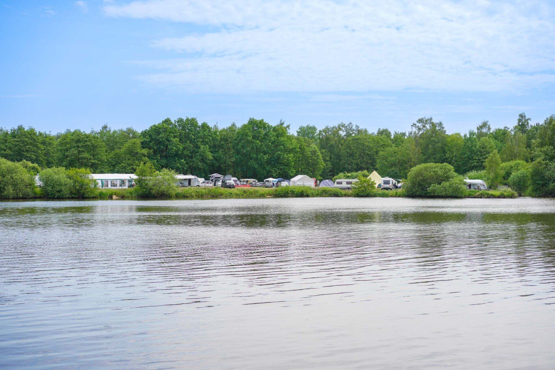 Seeblick mit Bäumen und entferntem Campingplatz unter blauem Himmel.