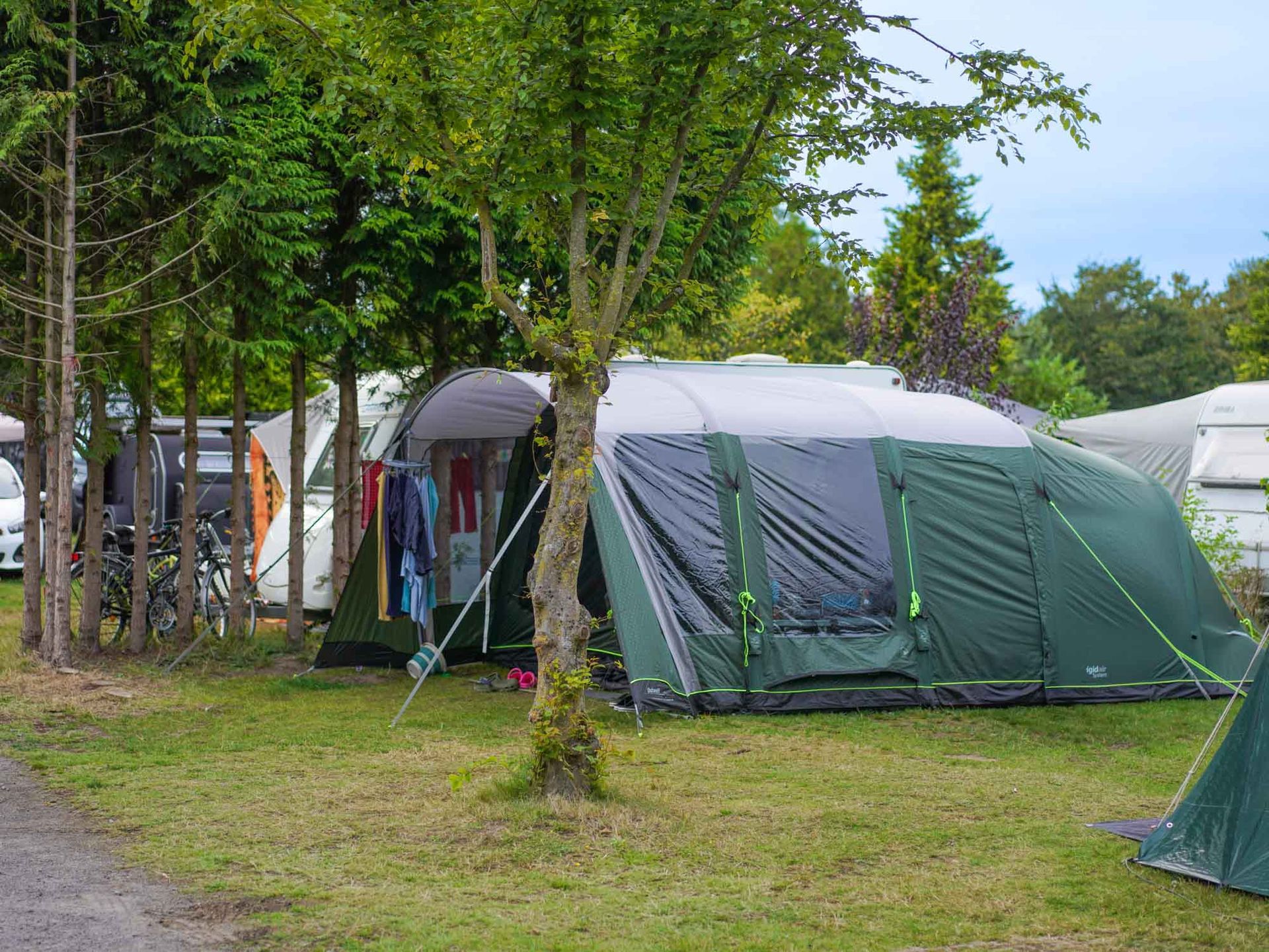 Grünes Zelt auf grasbewachsenem Campingplatz mit Bäumen, weiteren Zelten und Fahrzeug