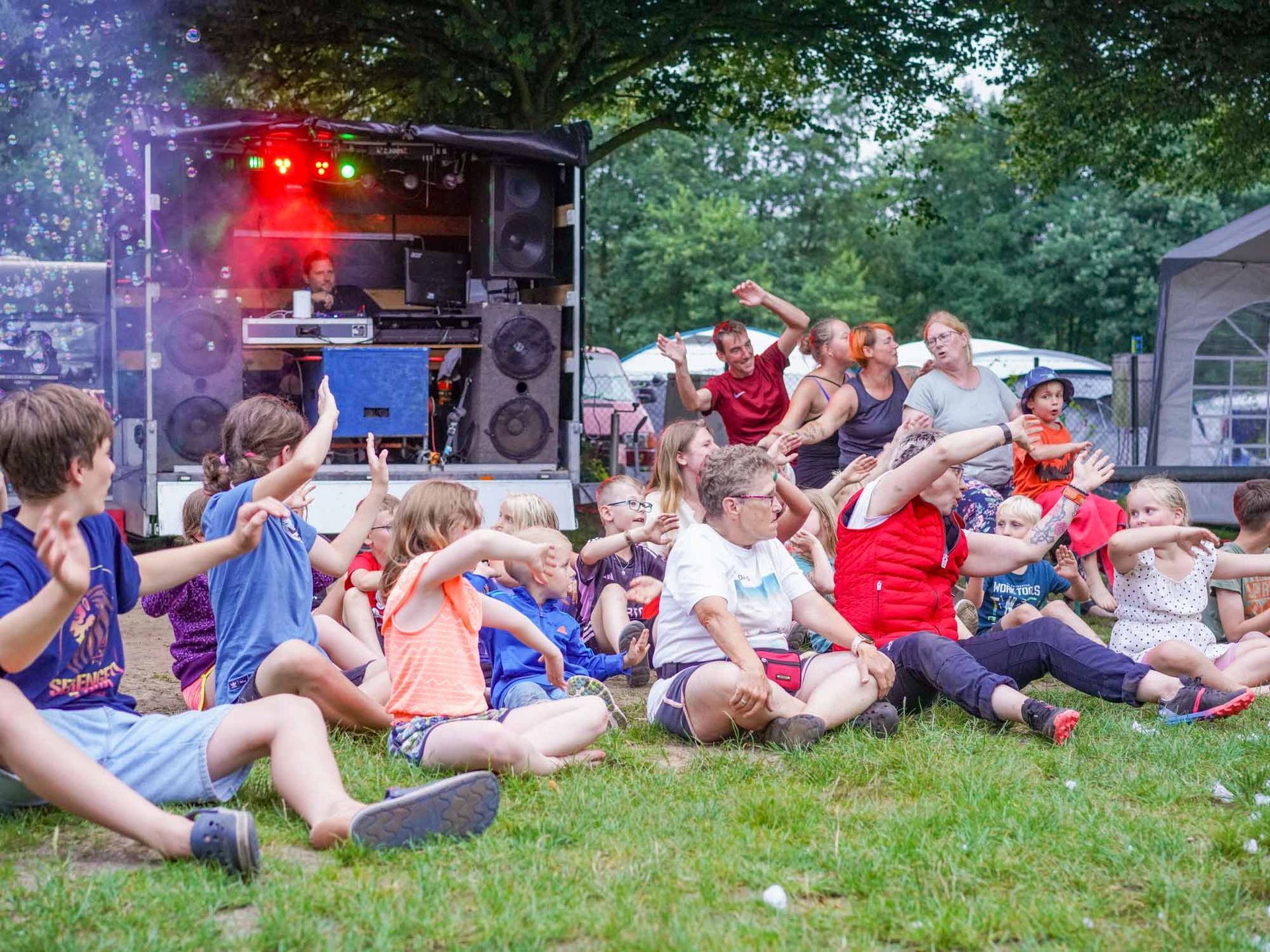 Kinder und Erwachsene tanzen auf einer Rasenfläche in der Nähe eines mobilen DJ-Pults mit Lautsprechern.